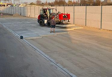 Betonplaten worden geplaatst met shovel op zandbed langs geluidswand voor terreinverharding